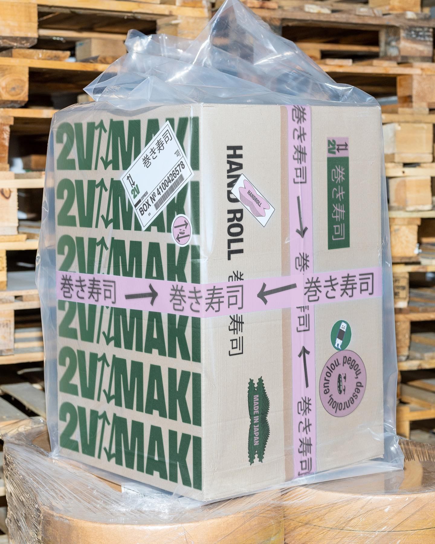 Caixa de papelão com a marca 2V Maki e escritas em japonês, embalada em plástico sobre um palete