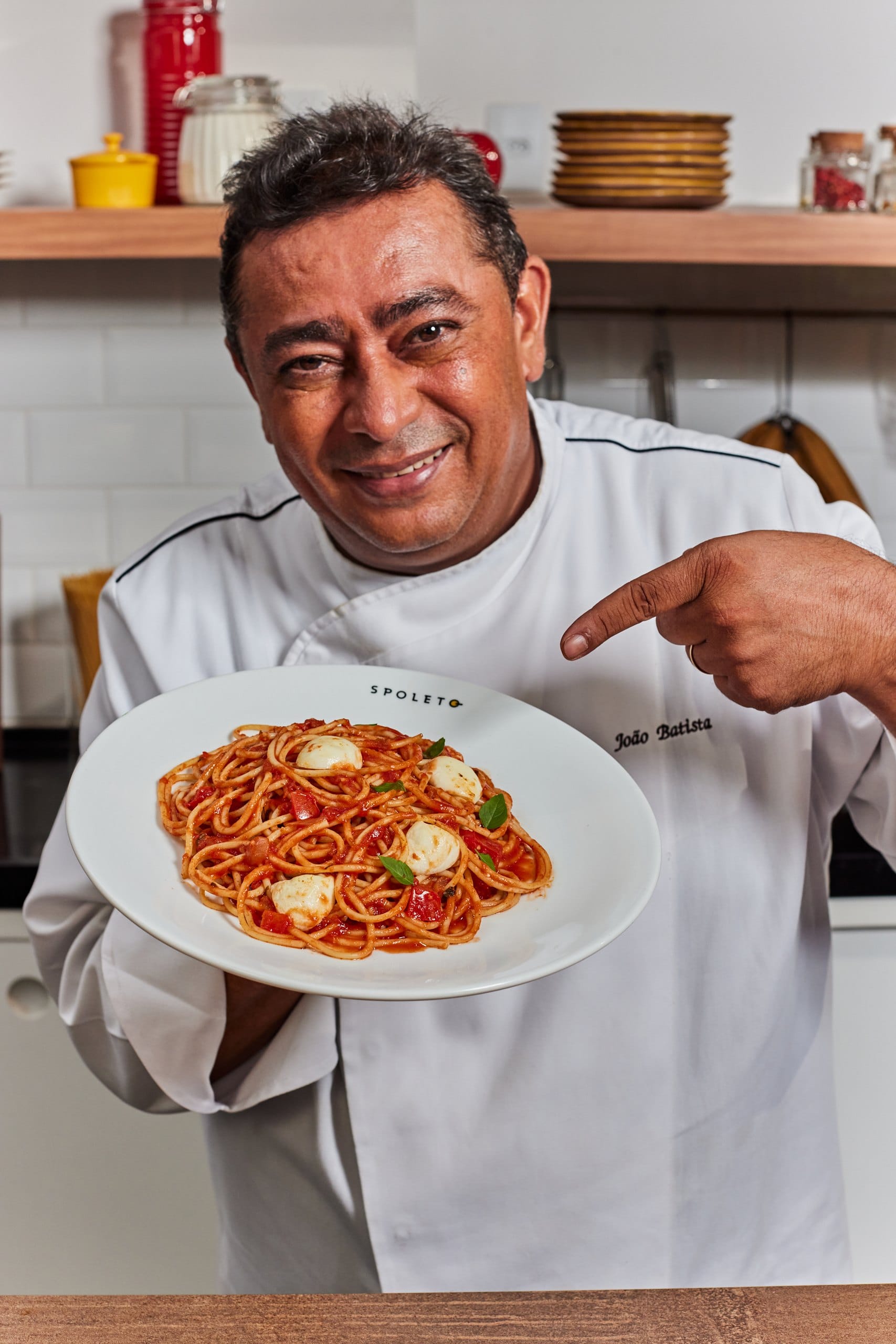 O chef João Batista sorri e aponta para um prato de espaguete com muçarela